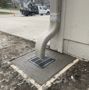 A downspout empties into a street drain set in freshly poured concrete near a driveway and parked utility trucks in the background.